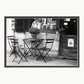 A black and white photograph showing three chairs and a table with a plant pot on top outside a famous bookshop in the Quartier Latin district in Paris. Shown with a double mount of black on the bottom and white on top