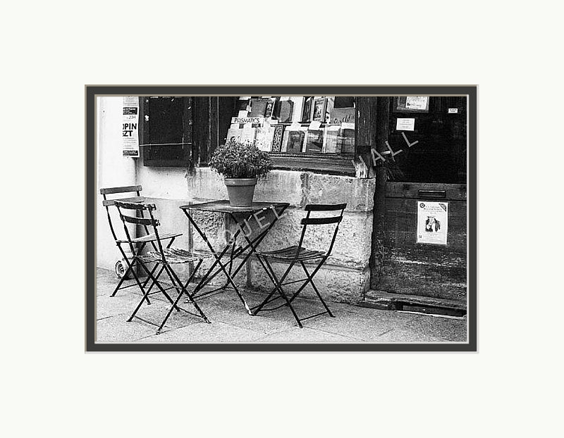 A black and white photograph showing three chairs and a table with a plant pot on top outside a famous bookshop in the Quartier Latin district in Paris. Shown with a double mount of black on the bottom and white on top