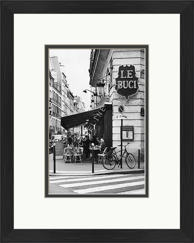 A black and white photograph of the corner of a Le Buci cafe in the Quartier Latin district in Paris. Shown in a black frame with a double mount of black on the bottom and white on top. 