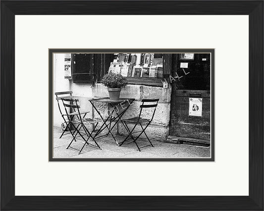 A black and white photograph showing three chairs and a table with a plant pot on top outside a famous bookshop in the Quartier Latin district in Paris. Shown in a black frame with a double mount of black on the bottom and white on top